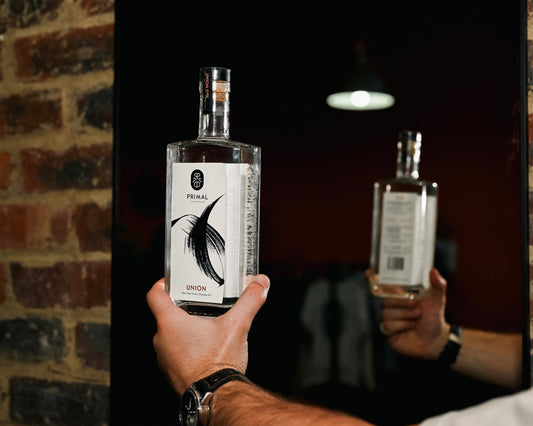Two bottles of 'Primal Union' gin held by a person in front of a brick wall.