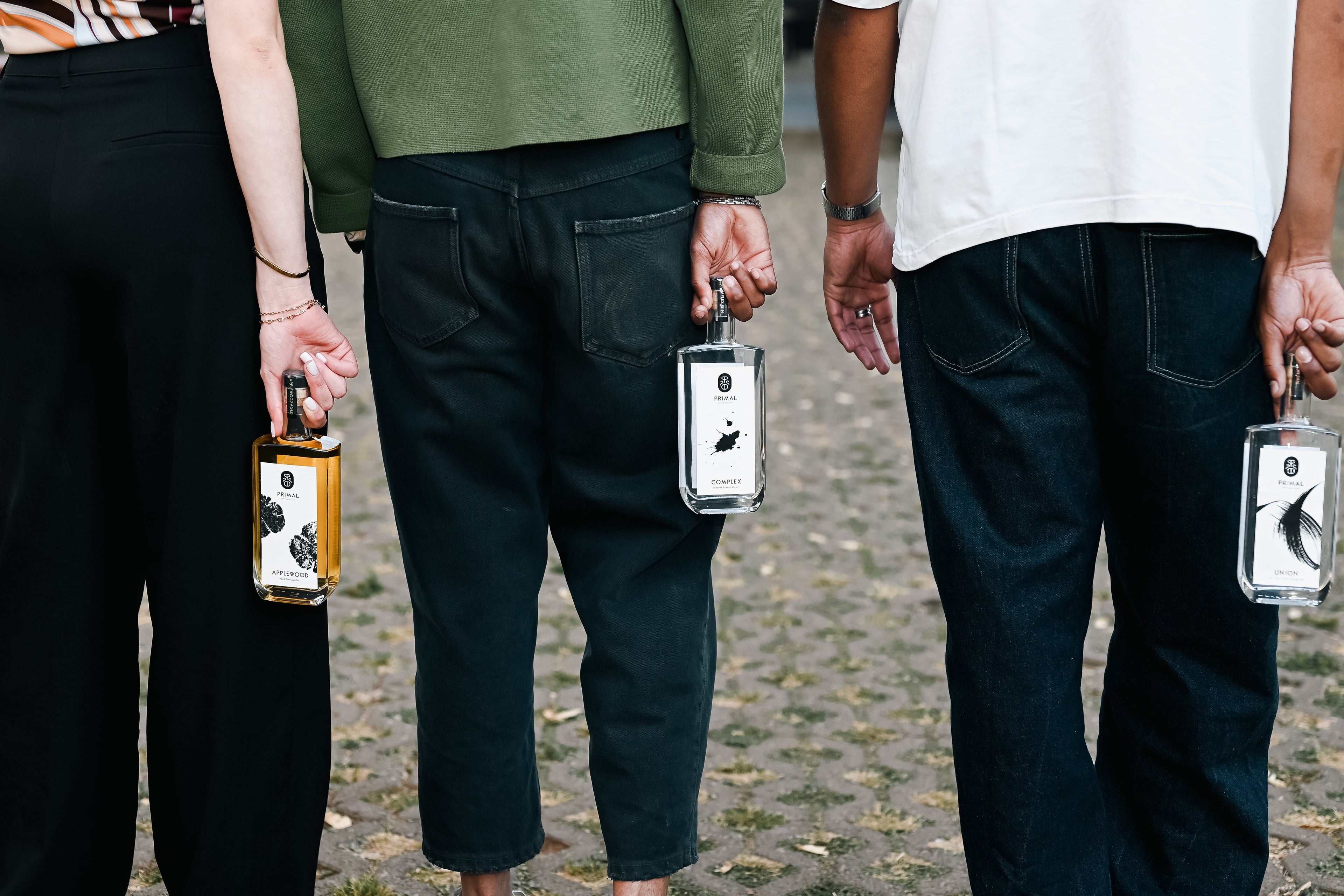 Three people standing outdoors holding bottles of Primal Gin. The people are shown from the waist down, wearing black, green, and white clothing on a cobblestone ground.